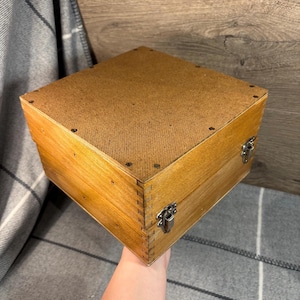 Puede incluir: Una caja de madera con tapa con bisagras y dos cierres metálicos. La caja está hecha de madera marrón claro y tiene un acabado natural. La tapa está forrada con una tela marrón.