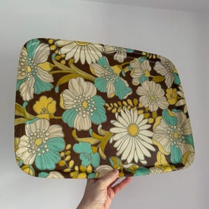 Peut inclure: Plateau rectangulaire avec un fond marron et un motif floral blanc, jaune et turquoise. Le plateau a des coins arrondis et est tenu par une main.