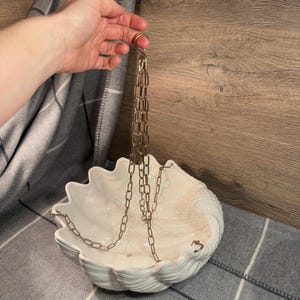 May include: White ceramic seashell planter with a gold chain for hanging. The planter is shaped like a clam shell and has a smooth, glossy finish.