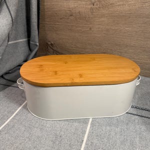 May include: An oval-shaped white metal storage bin featuring a smooth bamboo wood lid and small metal handles on each side. The container has a clean, solid white finish and a natural light brown wood grain texture on the top cover.