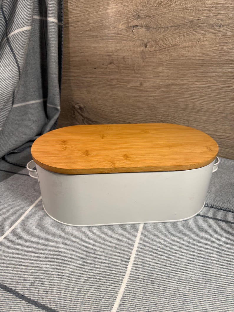 May include: An oval-shaped white metal storage bin featuring a smooth bamboo wood lid and small metal handles on each side. The container has a clean, solid white finish and a natural light brown wood grain texture on the top cover.