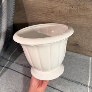May include: A white ceramic planter with a glossy finish, featuring a wide, rounded rim and a fluted, vertical ribbed body that tapers down to a circular pedestal base. The interior is smooth and hollow, showing a clean, white, reflective surface.