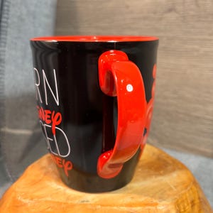 Peut inclure: Mug en c&eacute;ramique noire avec int&eacute;rieur et anse rouges. Le mug pr&eacute;sente un texte blanc qui se lit "BORN, RAISED, DISNEY, ED". La poign&eacute;e est de couleur rouge vif.