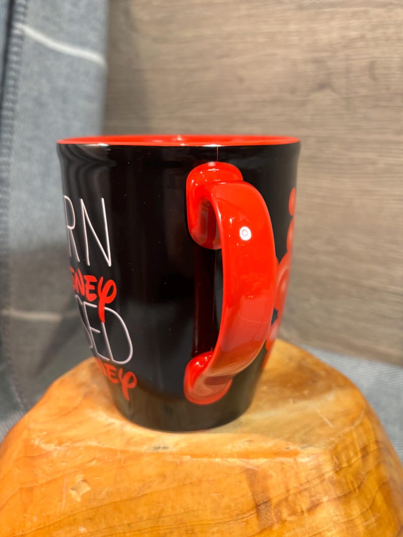 Peut inclure: Mug en c&eacute;ramique noire avec int&eacute;rieur et anse rouges. Le mug pr&eacute;sente un texte blanc qui se lit "BORN, RAISED, DISNEY, ED". La poign&eacute;e est de couleur rouge vif.