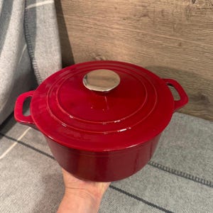 May include: A red cast iron Dutch oven with a silver knob on the lid. The pot has two handles and is sitting on a gray surface. The pot is a vibrant red color.