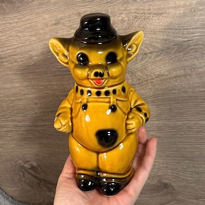 May include: A ceramic pig figurine featuring a glossy yellow finish with dark brown accents on the ears, hat, and clothing details. The pig is depicted wearing a hat and overalls, with a small red mouth and black painted eyes, nose, and buttons.