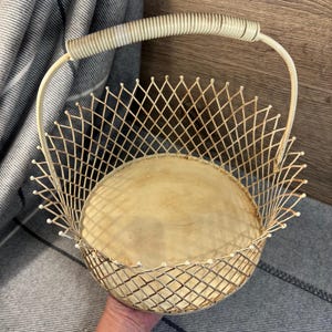 May include: Cream-coloured wire basket with a handle. The basket has a diamond-patterned wire design and a round, light-coloured base. The handle is wrapped with a textured material. The basket is likely vintage or antique.