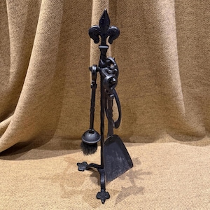 Puede incluir: Juego de herramientas para chimenea de hierro forjado negro. El juego incluye un atizador, un cepillo, una pala y un soporte. El soporte está adornado con diseños de flor de lis y volutas. Las herramientas están dispuestas verticalmente.