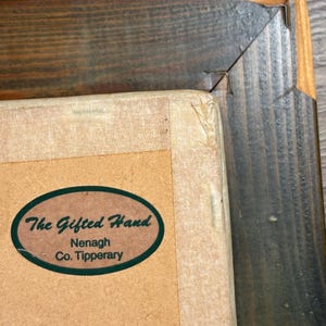 Puede incluir: Primer plano de un marco de imagen de madera marr&oacute;n con una pegatina ovalada verde que dice "The Gifted Hand Nenagh Co. Tipperary".