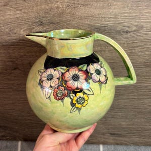 Może przedstawiać: Zielony dzbanek ceramiczny z czarną obwódką i kwiatowym wzorem. Kwiaty są różowe, żółte i białe z czarnymi konturami. Dzbanek ma złotą obwódkę i uchwyt.