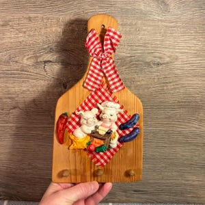 May include: Wooden kitchen wall decor with a decorative design. The decor features a red and white checkered bow, two pig figurines, a red chili pepper, and a small wooden box with vegetables. The decor has three small wooden pegs.