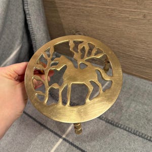 Może przedstawiać: Okrągły, złoty podkładka pod garnek z wyciętym wzorem przedstawiającym sylwetkę jelenia otoczoną stylizowanymi drzewami. Podkładka ma mały uchwyt i jest przeznaczona do ochrony powierzchni przed gorącem.