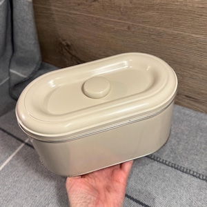 May include: A cream-coloured, oval bread bin with a matching lid and a small, round knob. The bin has a simple, classic design with a thin, silver trim around the lid and base. The bin is made of metal.
