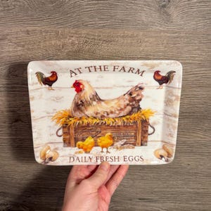 Puede incluir: Bandeja rectangular de cerámica con temática de granja. La bandeja presenta una gallina, pollitos y gallos, con el texto "AT THE FARM" y "DAILY FRESH EGGS". La bandeja está sujeta por una mano.
