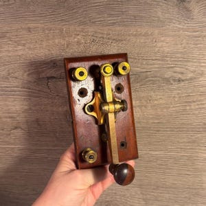 May include: A vintage telegraph key mounted on a rectangular dark brown wooden base. The device features a brass lever arm with a rounded dark brown knob at the end, along with several brass binding posts and screws for electrical connections.