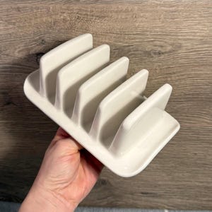 Puede incluir: Un tostador de cerámica blanca con cinco ranuras. El soporte tiene una base rectangular y divisores curvos. El acabado brillante refleja la luz, resaltando el diseño liso y limpio. Diseñado para sostener la tostada en posición vertical.