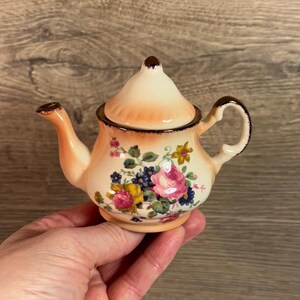 Puede incluir: Una tetera pequeña de color crema con un diseño floral. La tetera tiene una tapa y un asa. El diseño floral presenta flores rosas, amarillas y azules.