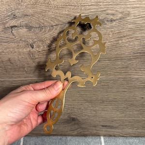 May include: A gold-colored metal trivet with an ornate, openwork design. The trivet features a handle with a circular opening at the end. The decorative piece is designed to protect surfaces from hot cookware.