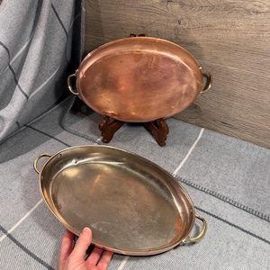 May include: Two oval copper serving dishes with brass handles. One dish is positioned upright on a wooden stand, displaying its polished copper exterior. The second dish is held in the foreground, showing its metallic interior surface.