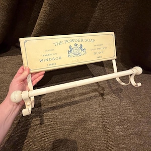 May include: A cream-colored metal towel rack featuring a rectangular sign with blue text that reads THE POWDER SOAP, GENUINE FAMILY WINDSOR LONDON, EMULLIENT OLD BROWN SOAP PARIS, and a central blue crest illustration with two horses.
