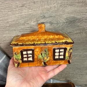 Puede incluir: Una caja de cerámica con forma de cabaña con techo de paja, paredes de ladrillo marrón y dos ventanas. La caja está decorada con hojas y enredaderas pintadas de verde y amarillo.