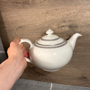 Könnte beinhalten: Weiße Porzellan-Teekanne mit silbernem Rand um Deckel und Korpus. Die Teekanne hat einen runden Korpus und einen Ausguss auf der rechten Seite.