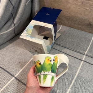 May include: White ceramic mug featuring a vibrant illustration of three green parrots with yellow and orange accents perched on a branch. The mug has a curved handle and is accompanied by its original box.
