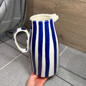 May include: A ceramic pitcher featuring a white base with vertical dark blue stripes and a glossy crackle glaze finish. The vessel has a curved handle on the side and a wide, moulded pouring spout at the top rim, showcasing a hand-painted aesthetic.