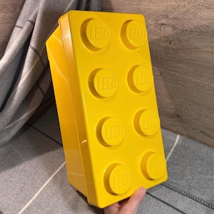 May include: Bright yellow LEGO brick-shaped storage container. The container has eight raised circular studs, each with the LEGO logo. The container is held against a gray and white patterned background.