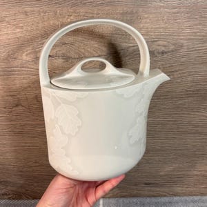 May include: White ceramic teapot with a leaf design. The teapot has a lid and a handle.