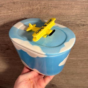 Könnte beinhalten: Ein Keramikbehälter mit Deckel, bemalt mit einem Himmel- und Wolkenmuster. Ein gelbes Flugzeug mit dem Text "Utterly Butterfly" sitzt auf dem Deckel. Der Behälter wird in einer Hand gehalten.