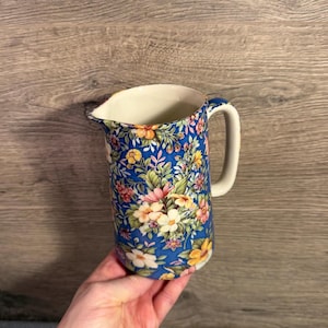 May include: A blue ceramic pitcher with a floral pattern. The pitcher has a white handle and a spout. The floral design includes pink, yellow, and white flowers with green leaves. The background is a wooden surface.
