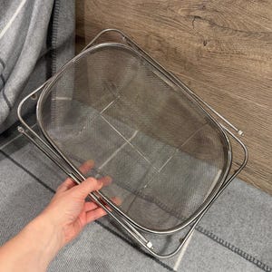 以下が含まれることがあります： 長方形のステンレス鋼メッシュザル。折りたたみ式のハンドルが付いています。食品から液体を排出するように設計されています。メッシュは効率的な水の除去を可能にします。グレーと白のチェック柄の毛布の上に置かれています。