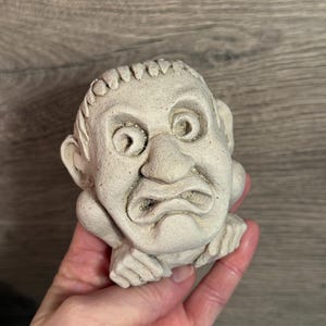 May include: A small, off-white, sculpted gargoyle head with a furrowed brow and a downturned mouth. The sculpture has detailed features, including visible ears and hands supporting the chin. The piece appears to be made of a textured, stone-like material.