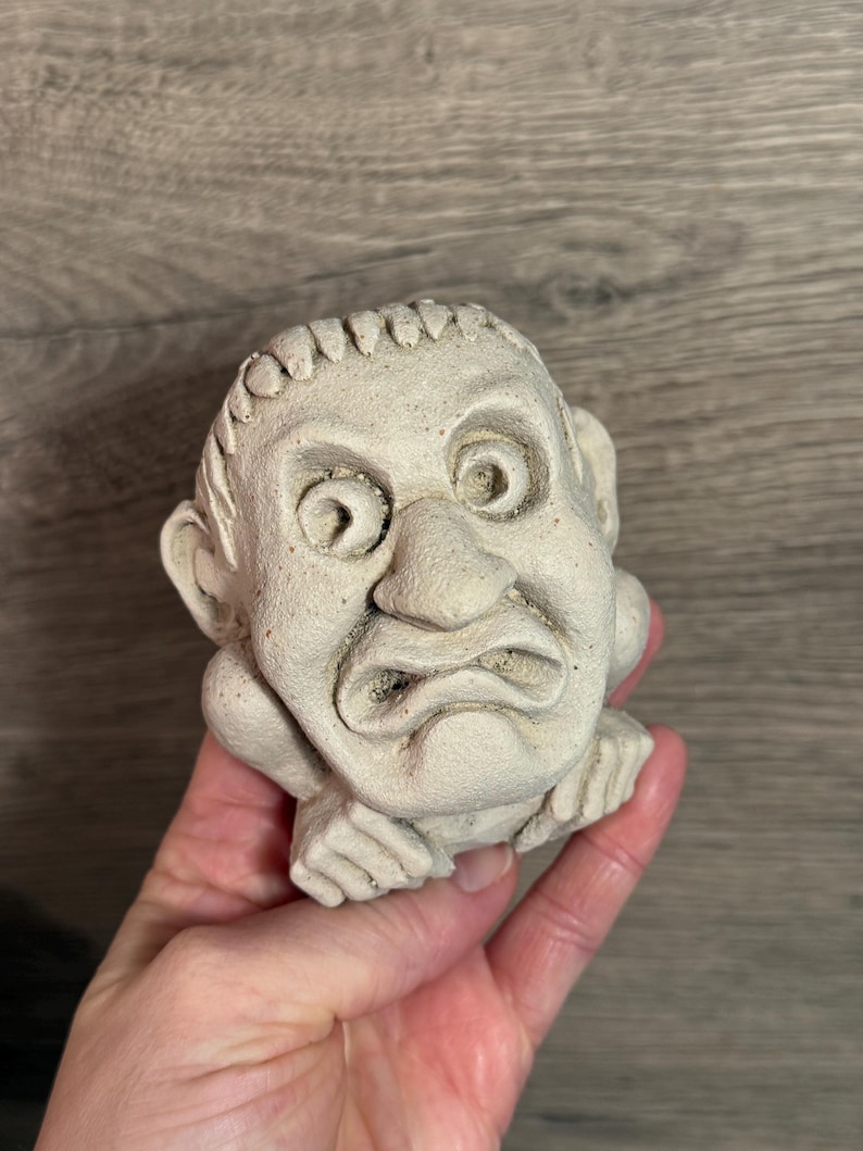 May include: A small, off-white, sculpted gargoyle head with a furrowed brow and a downturned mouth. The sculpture has detailed features, including visible ears and hands supporting the chin. The piece appears to be made of a textured, stone-like material.