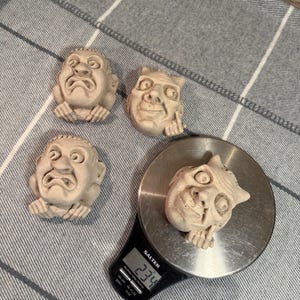 May include: Four small, beige-colored, sculpted face figurines. One figurine is on a digital scale, which reads 234. The figurines have exaggerated features and a distressed appearance. The scale is black and silver.