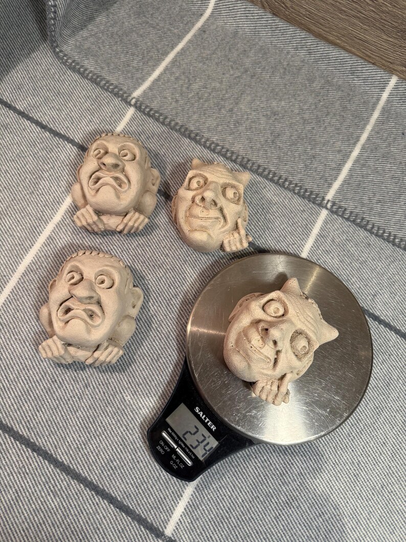May include: Four small, beige-colored, sculpted face figurines. One figurine is on a digital scale, which reads 234. The figurines have exaggerated features and a distressed appearance. The scale is black and silver.