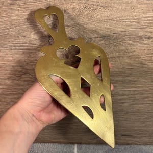 Op de afbeelding: Een gouden metalen hartvormig serveerbestek met een ingewikkeld uitgesneden ontwerp. Het bestek heeft een handvat met een hartvormige opening.