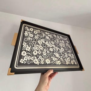 May include: A rectangular serving tray with a black frame and a floral design. The tray features a black background with a white floral pattern. It has gold-colored corner accents and wooden handles.