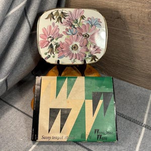 May include: A vintage floral teapot stand made by Worcester Ware. The stand is made of metal and has a green and black geometric design. The stand is in its original box, which says "Savoy teapot stand".