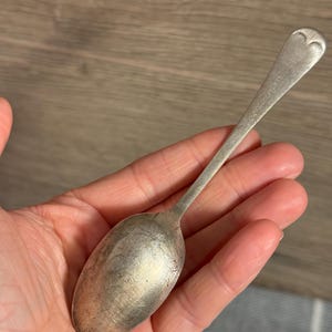 Op de afbeelding: Een zilveren lepel met een hartvormig ontwerp aan het uiteinde van het handvat. De lepel heeft een lange, slanke steel en een afgeronde kom. De lepel wordt in de hand van een persoon gehouden.