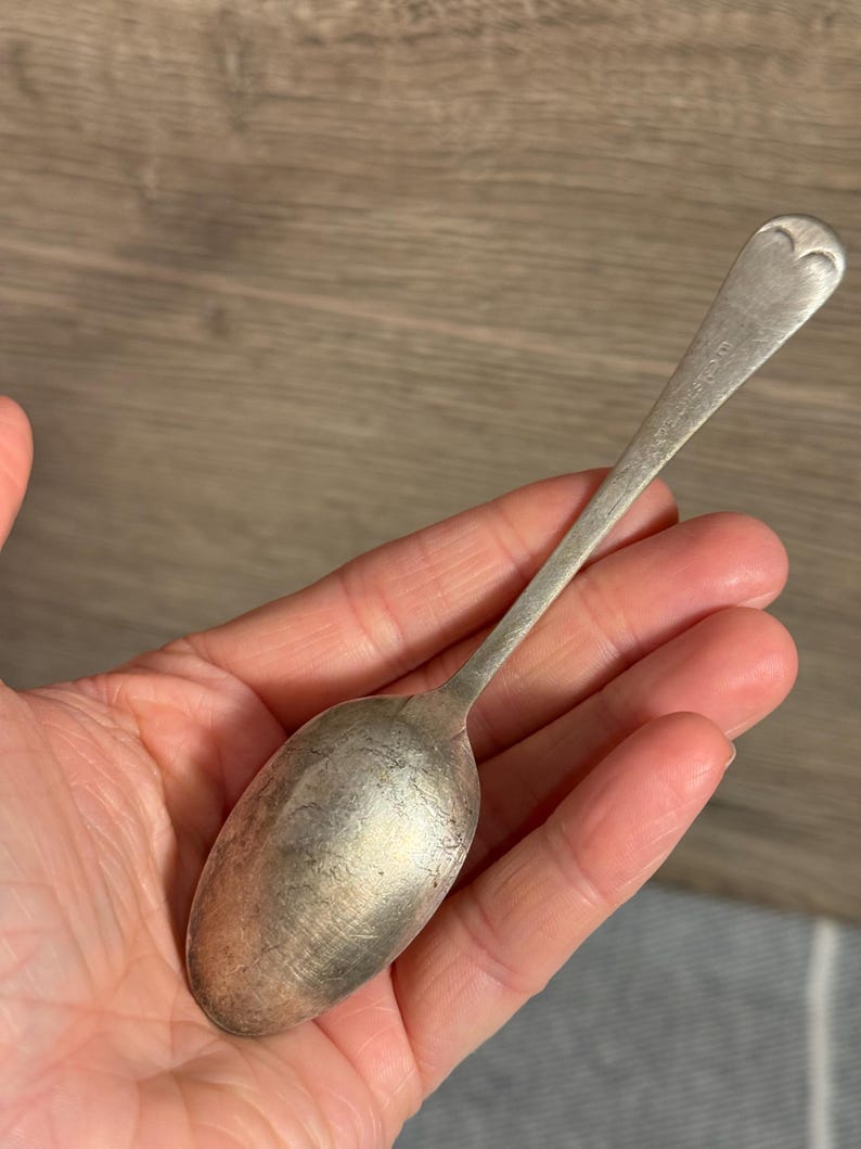 Op de afbeelding: Een zilveren lepel met een hartvormig ontwerp aan het uiteinde van het handvat. De lepel heeft een lange, slanke steel en een afgeronde kom. De lepel wordt in de hand van een persoon gehouden.