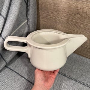 May include: A cream-coloured ceramic jug with a handle and a spout. The jug has a rounded body and a wide opening. The handle is a loop shape. The jug is held in a hand.