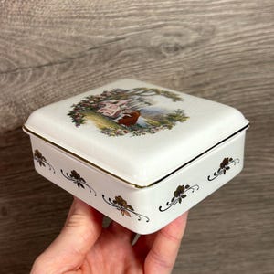 Op de afbeelding: Een vierkante, witte keramische sieradendoos met een deksel met een pastorale scène. De doos heeft gouden randen en bloemmotieven langs de zijkanten. Het deksel toont een stel onder een boom.