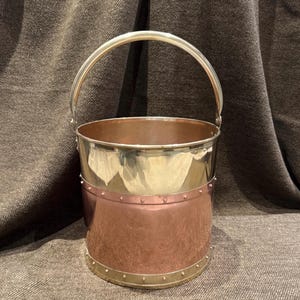 May include: A vintage metal bucket with a shiny silver handle and rim. The upper section is polished silver, the middle is copper, and the base is silver. The bucket has a cylindrical shape and is riveted with small studs.