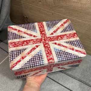 May include: Rectangular tin box featuring the Union Jack flag design. The flag's red and white stripes are overlaid on a blue and white checkered background. The box is held in a hand.