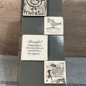 May include: A grey wooden box with a decorative design. The box has a series of square tiles with inspirational quotes and images of birds. The quotes include "Tweet!" "Beautiful things happen when you distance yourself from the negative." "Praise God for the rain, all blessings flow." and "I choose to be unstoppable. I am bigger than my concerns and worries. The strength of others inspire me daily. I focus on my goal. I trust my intuition and live a courageous life."