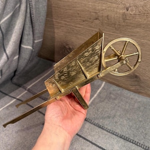 Könnte beinhalten: Eine Miniatur-Schubkarre aus Messing mit detailliertem Design. Die Schubkarre hat ein einzelnes Rad und zwei lange Griffe. Das Messing hat eine warme, goldene Farbe. Die Schubkarre ist ein dekorativer Artikel.