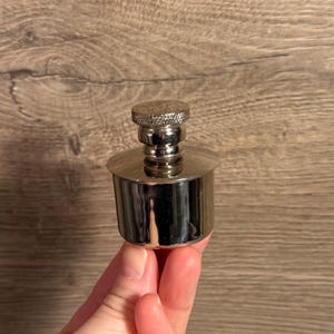 Puede incluir: Un tapón de botella plateado con una parte superior texturizada y una base cilíndrica. El tapón está hecho de metal brillante y tiene un diseño clásico. La imagen muestra un primer plano del tapón, destacando sus detalles.