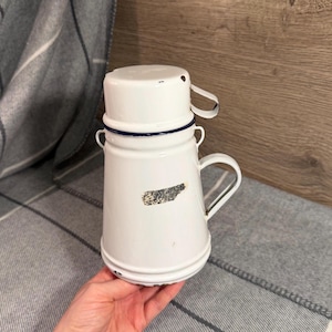 May include: White enamelware coffee pot with a lid and handle. The pot has a blue trim and a small handle on the lid. A faded label is visible on the side. The pot is held in a person's hand.